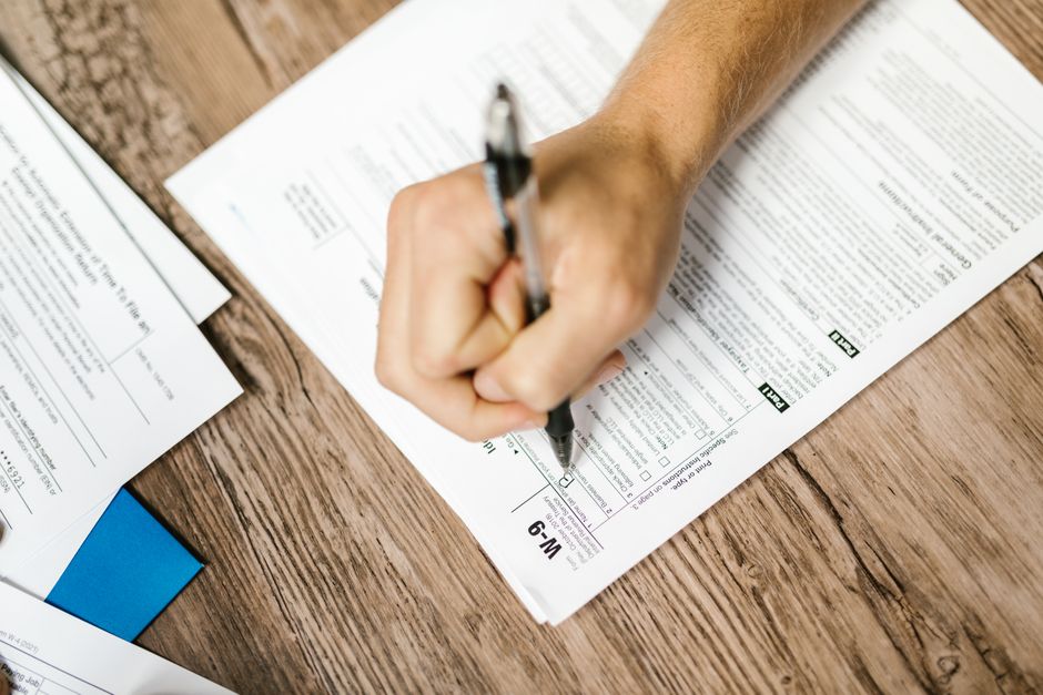 Hand writing on a W-9 tax form using a ballpoint pen on wooden desk.